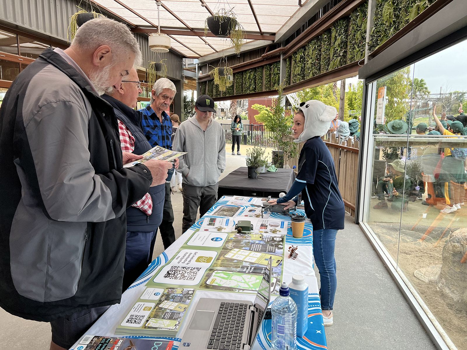 Four men look at brochures from a stall staffed by a girl in a koala hat