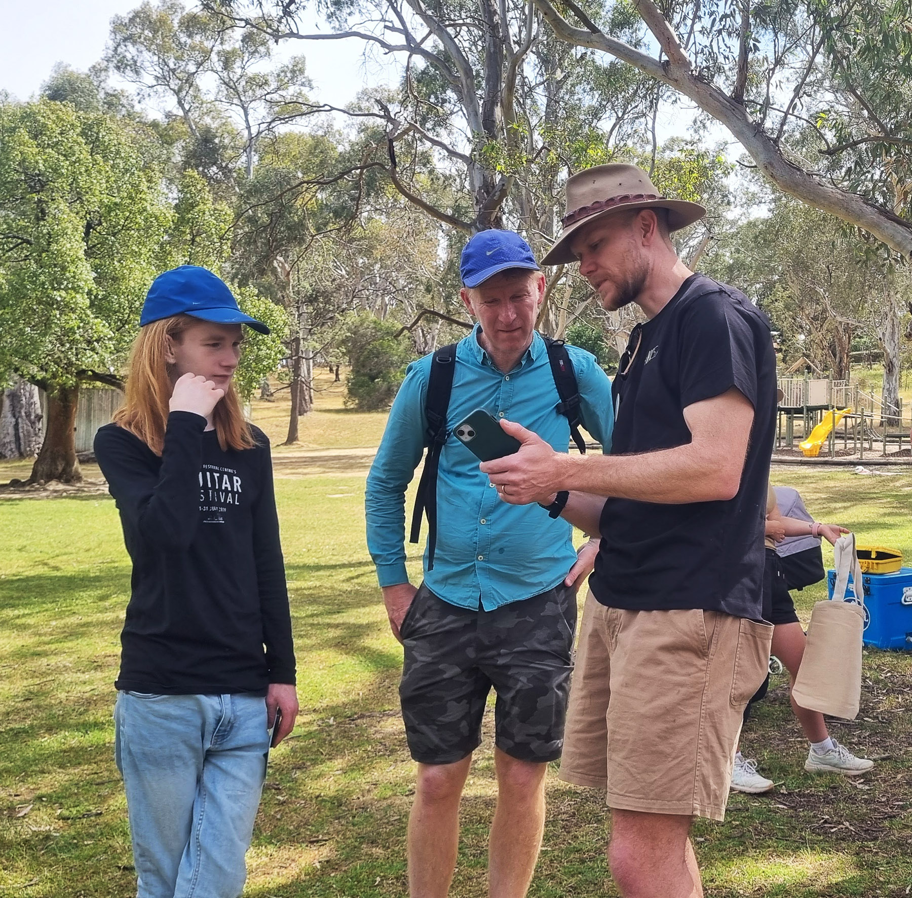 Three people talk and look at a phone in a parkland